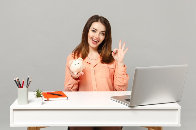 Una joven mujer divertida con ropa pastel que muestra un gesto OK sostiene un banco de dinero alcancía trabajando en el escritorio con una computadora portátil aislada en un fondo gris. Concepto de estilo de vida de carrera empresarial de logro. Simulacros de espacio de copia.