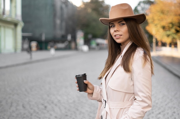 Joven mujer elegante de apariencia europea se para en la calle y bebe café de vajilla desechable.