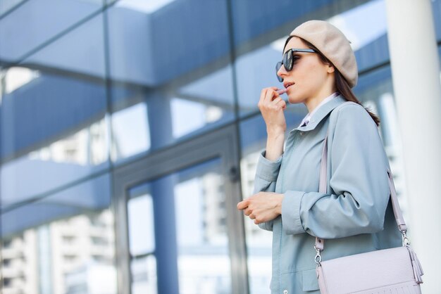 álbum Críticamente Centelleo Joven mujer elegante con gafas de sol y boina pinta sus labios contra el  fondo de las ventanas del centro de negocios estilo de vida de belleza de  moda | Foto Premium