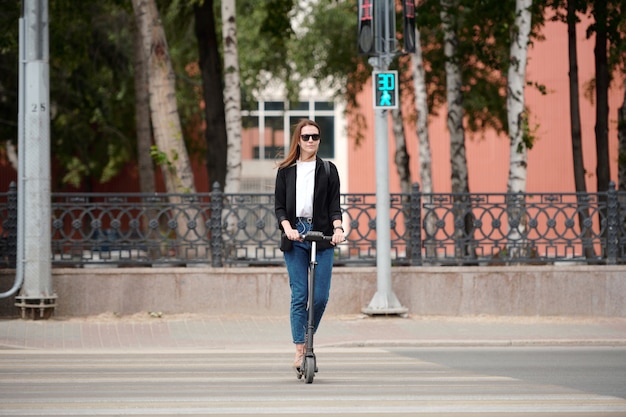 Joven mujer elegante con gafas de sol y ropa informal de pie en scooter eléctrico y bajando por el paso de peatones mientras las luces verdes están encendidas