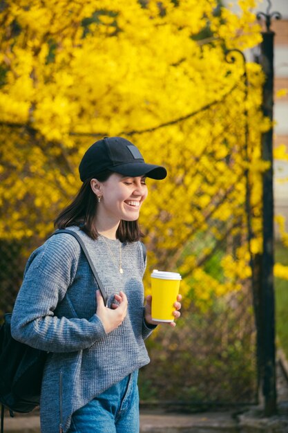 Joven mujer elegante con gorra bebiendo café al aire libre en un vaso de papel amarillo