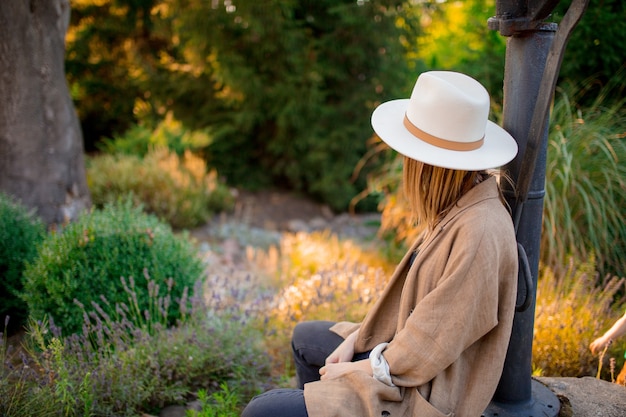 Joven mujer elegante con sombrero sentado cerca de flores de lavanda en un jardín.