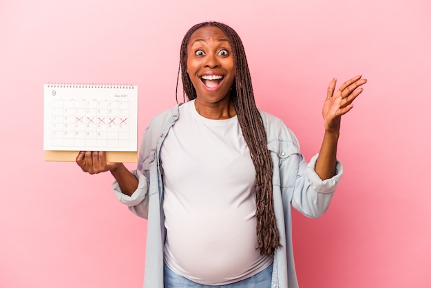 Joven mujer embarazada afroamericana con calendario aislado sobre fondo rosa recibiendo una agradable sorpresa, emocionada y levantando las manos.