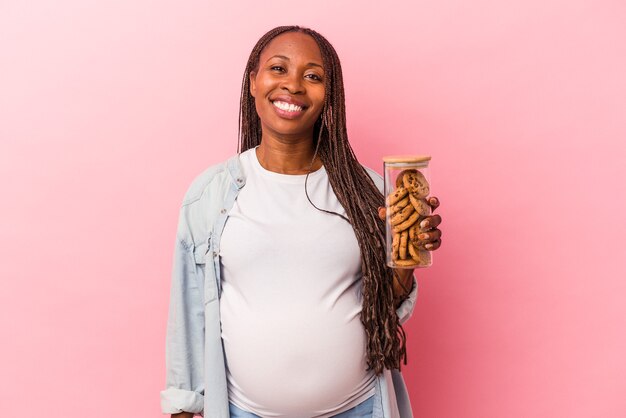 Joven mujer embarazada afroamericana con galletas aisladas sobre fondo rosa feliz, sonriente y alegre.