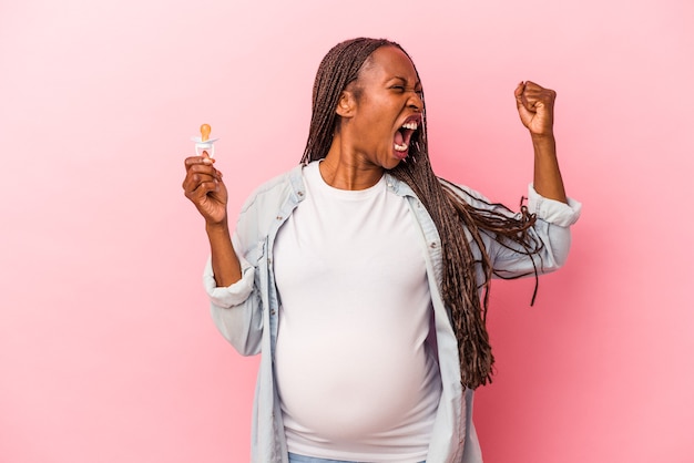 Joven mujer embarazada afroamericana sosteniendo chupete aislado sobre fondo rosa levantando el puño después de una victoria, concepto ganador.