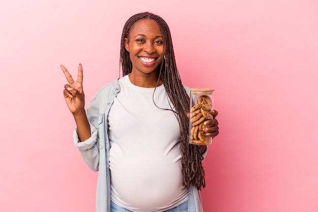 Joven mujer embarazada afroamericana sosteniendo galletas aisladas sobre fondo rosa mostrando el número dos con los dedos.