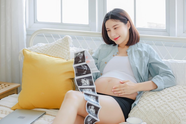 Joven mujer embarazada asiática feliz es y sosteniendo ultrasonido imagen de escaneo 4D tocando su vientre plan de familia embarazo maternidad personas y expectativa niño y maternidad concepto de atención prenatal