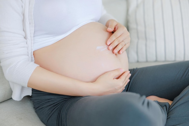 Foto joven mujer embarazada asiática feliz está sentada en la cama y aplicar crema o suero en el vientre plan de familia embarazo maternidad personas y expectativa niño y maternidad concepto de atención prenatal