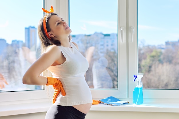 Joven mujer embarazada cansada en guantes con trapo y ventana de lavado de detergente