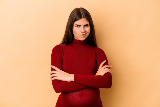 Joven mujer embarazada caucásica aislada en la pared beige con el ceño fruncido en disgusto, mantiene los brazos cruzados.