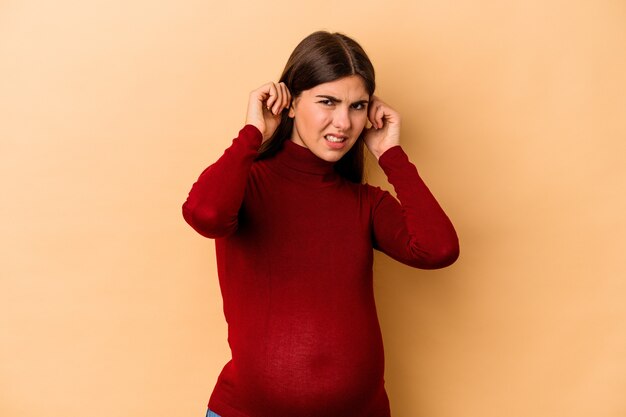 Joven mujer embarazada caucásica aislada en la pared beige que cubre las orejas con las manos