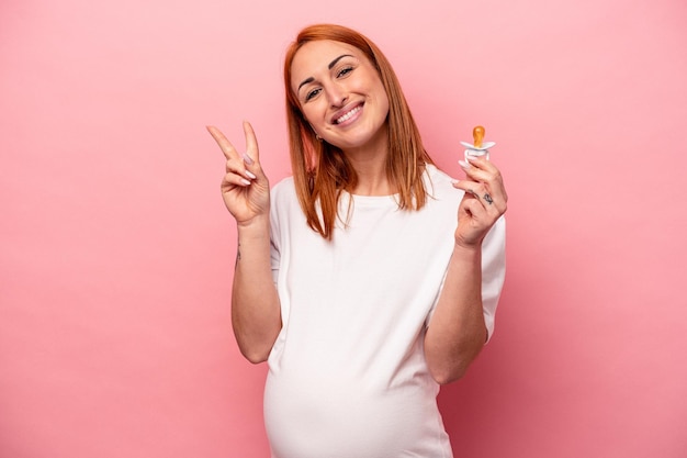 Foto joven mujer embarazada caucásica sosteniendo un chupete aislado en un fondo rosado alegre y despreocupada mostrando un símbolo de paz con los dedos