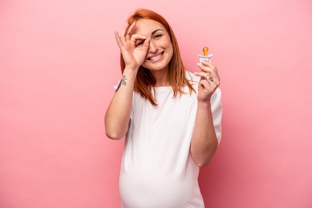 Joven mujer embarazada caucásica sosteniendo chupete aislado sobre fondo rosa emocionado manteniendo el gesto de ok en el ojo