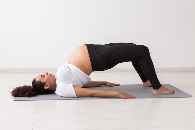 Joven mujer embarazada flexible haciendo gimnasia sobre una alfombra en el suelo de la pared blanca. El concepto de preparar el cuerpo para un parto fácil.