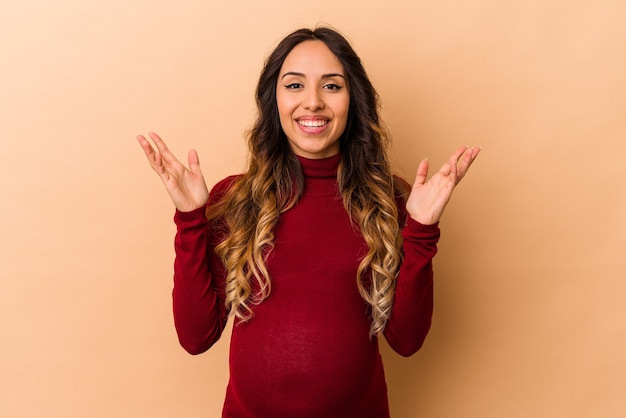 Joven mujer embarazada mexicana aislada sobre fondo beige recibiendo una agradable sorpresa, emocionada y levantando las manos.