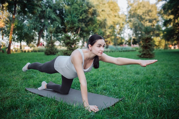 Joven mujer embarazada de pie sobre las rodillas y hacer ejercicio en compañero de yoga en el parque. Ella estira las piernas y los brazos. Mujer concentrada y seria mirar hacia adelante.