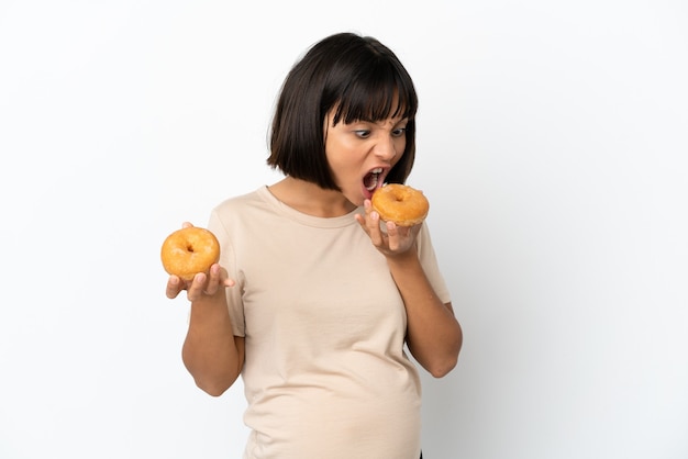 Joven mujer embarazada de raza mixta aislada sobre fondo blanco comiendo una rosquilla