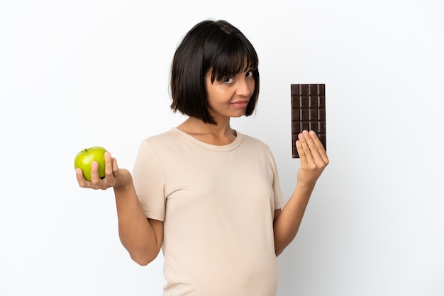 Joven mujer embarazada de raza mixta aislada sobre fondo blanco tomando una tableta de chocolate en una mano y una manzana en la otra