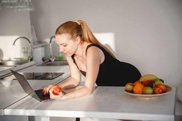 Joven mujer embarazada rubia vestida de negro en la cocina blanca comiendo fruta y mirando portátil. Foto de alta calidad