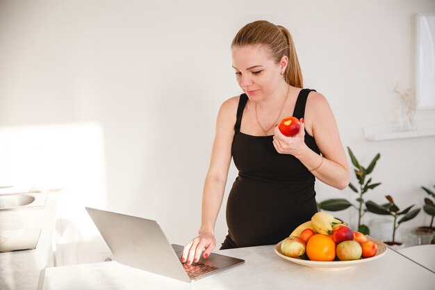 Joven mujer embarazada rubia en vestido negro en la cocina blanca comiendo fruta y mirando portátil