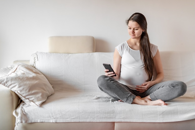 Joven mujer embarazada sentada en el sofá blanco con teléfono inteligente y manteniendo la mano sobre el vientre