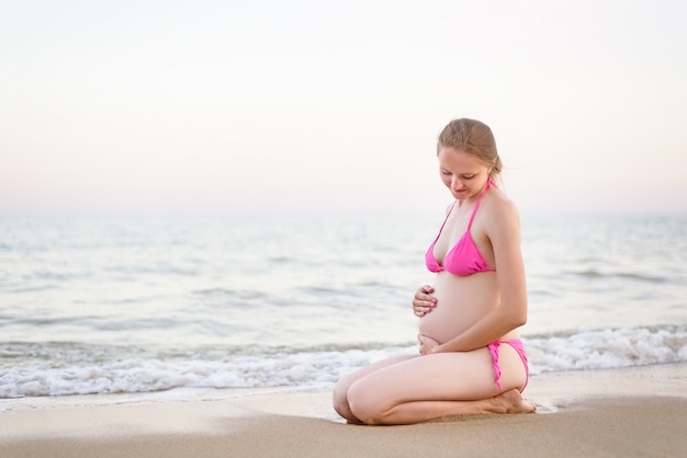 Joven mujer embarazada en traje de baño sentada en la playa y abrazando su vientre. Hora de verano antes del atardecer