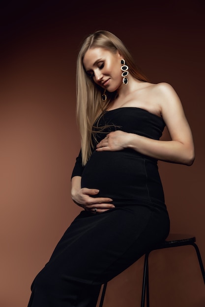 Una joven mujer embarazada en un vestido está de pie sobre un fondo marrón. Foto tomada en un estudio fotográfico.