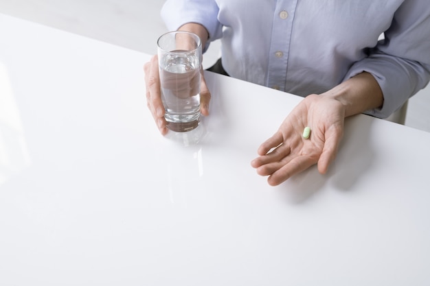 Joven mujer enferma o empresaria en camisa sosteniendo un vaso de agua y una pastilla mientras está sentado junto al escritorio