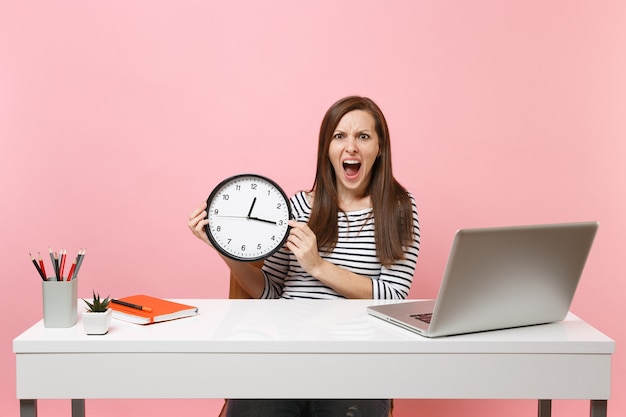 Joven mujer enojada sosteniendo un reloj despertador redondo gritando mientras trabaja sentado en la oficina con un ordenador portátil aislado sobre fondo rosa pastel. Concepto de carrera empresarial de logro. Copie el espacio. Se acaba el tiempo.