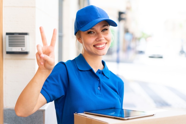 Foto joven mujer de entrega al aire libre sosteniendo cajas y una tableta y haciendo el gesto de la victoria