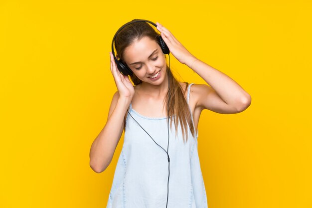 Joven mujer escuchando música sobre pared amarilla aislada cantando