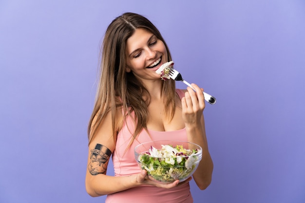 Joven mujer eslovaca aislada en púrpura sosteniendo un plato de ensalada y mirándolo con expresión feliz