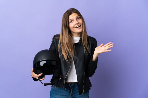 Joven mujer eslovaca sosteniendo un casco de motocicleta aislado en la pared púrpura con expresión facial conmocionada