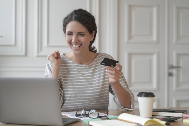 Joven mujer española feliz tiene tarjeta de crédito y se siente emocionada mientras hace compras en línea en una computadora portátil