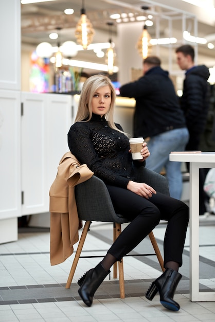 Una joven mujer está sentada en la cafetería de un centro comercial, chatea por teléfono móvil y sostiene una taza de café de papel en la mano.
