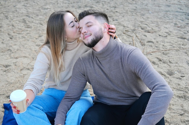 Un joven y una mujer están sentados en la orilla del lago bebiendo café y abrazándose. La chica abraza al chico y lo besa en la frente.