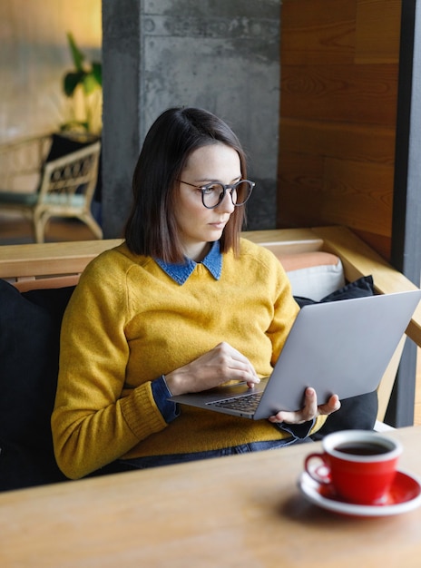 Una joven mujer europea caucásica en una camisa de mezclilla azul y gafas está trabajando en una computadora portátil