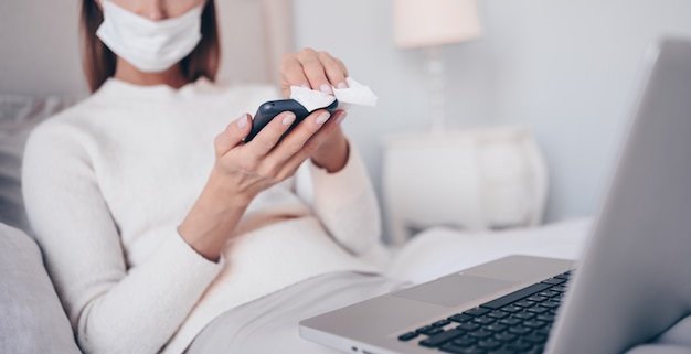 Joven mujer europea en mascarilla en el dormitorio con la computadora portátil durante el aislamiento de coronavirus