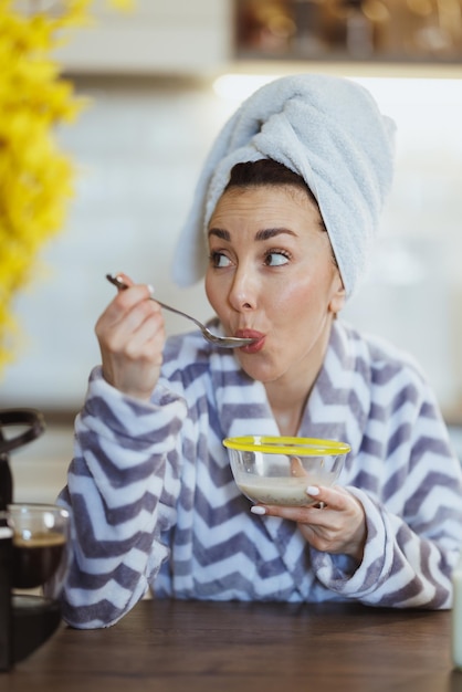 Joven mujer feliz en albornoz comiendo cereales para desayunar en la cocina de casa.