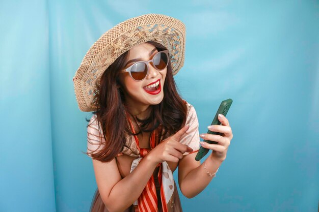 Joven mujer feliz asiática señalando y sonriendo a su teléfono inteligente aislado por un fondo azul