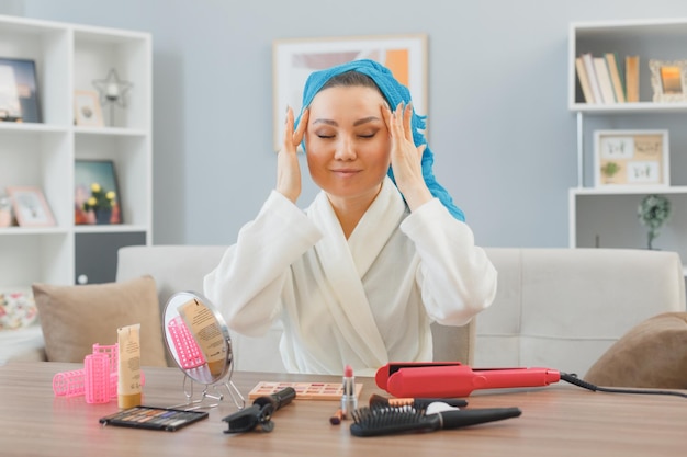 Joven mujer feliz asiática con una toalla en la cabeza sentada en el tocador en el interior de la casa mirando en el espejo haciendo un masaje facial concepto de belleza y cosmética facial