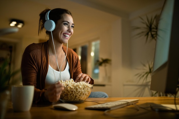 Joven mujer feliz con auriculares viendo películas en una computadora y comiendo palomitas de maíz por la noche en casa