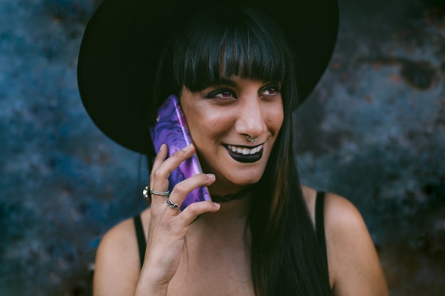 Joven mujer feliz bruja de Halloween con teléfono inteligente. Bella dama bruja con boca negra en la oscuridad, con un sombrero de bruja.