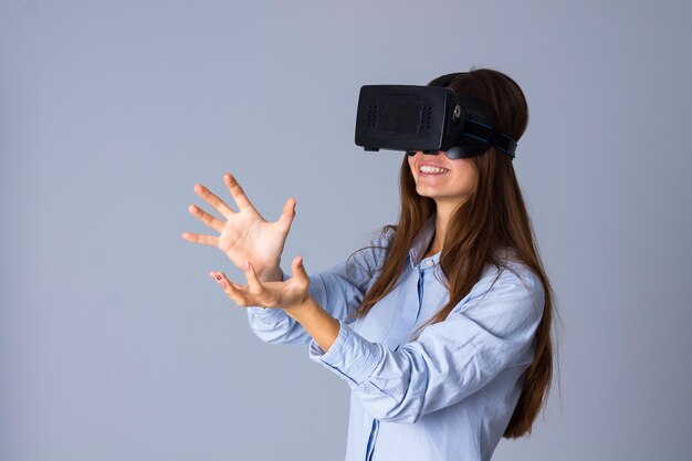 Joven mujer feliz con camisa azul usando gafas VR sobre fondo gris en el estudio