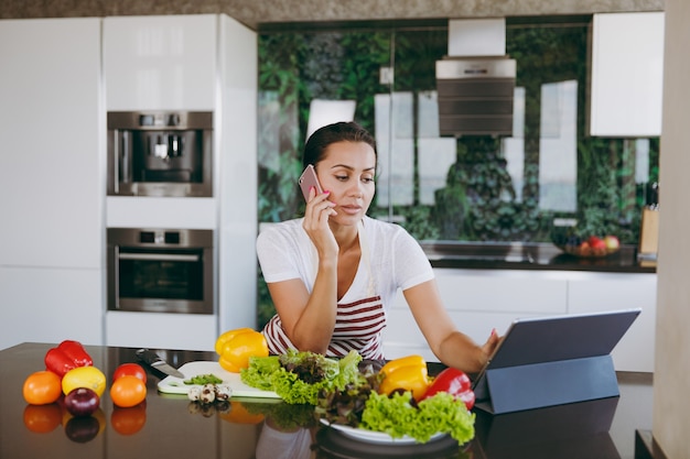 La joven mujer feliz en delantal hablando por teléfono móvil y mirando la receta en la computadora portátil en la cocina