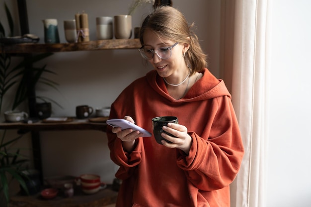 Joven mujer feliz dueña de una tienda de cerámica usando teléfono y bebiendo té charlando con clientes en línea