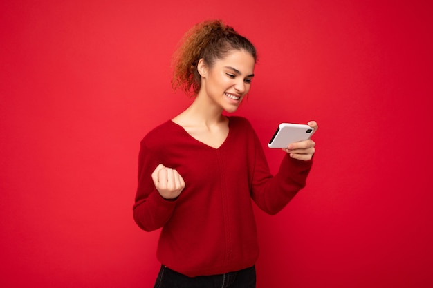 Joven mujer feliz emocional vistiendo suéter rojo oscuro aislado sobre fondo rojo con smartphone