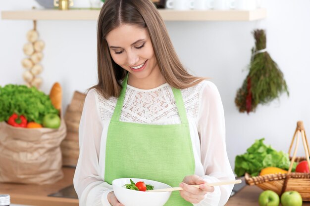 Joven mujer feliz está cocinando o comiendo ensalada fresca en la cocina Concepto de comida y salud