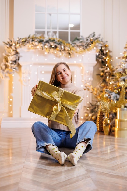 Una joven mujer feliz está sentada con un árbol de Navidad de regalo de Año Nuevo y una chimenea con guirnaldas festivas Estado de ánimo de Año Nuevo