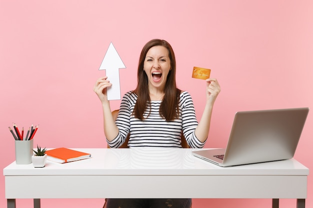 Joven mujer feliz gritando sosteniendo la flecha hacia arriba, tarjeta de crédito sentada y trabajando en la oficina con computadora portátil contemporánea